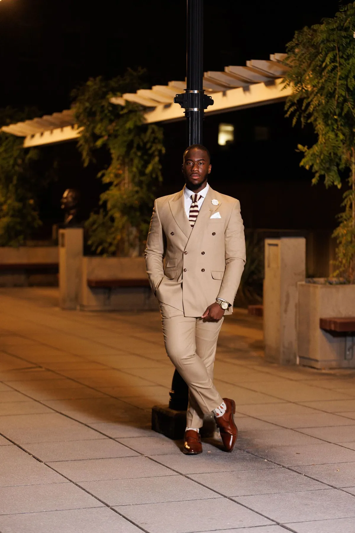 Adel Bebe in a tan double-breasted suit and knit tie, leaning against a lamppost in an evening courtyard