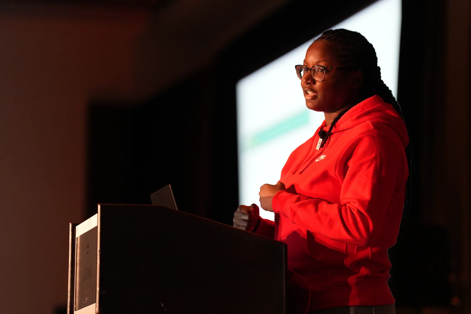 Danielle Lewis presenting on stage, lit in red, with a slide projected behind her