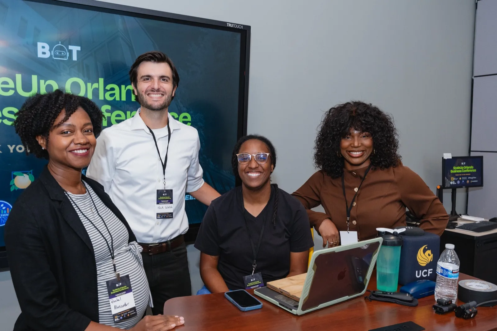 Four people at the check-in desk of the ScaleUp Orlando Business Conference, in front of a screen carrying the BOT logo