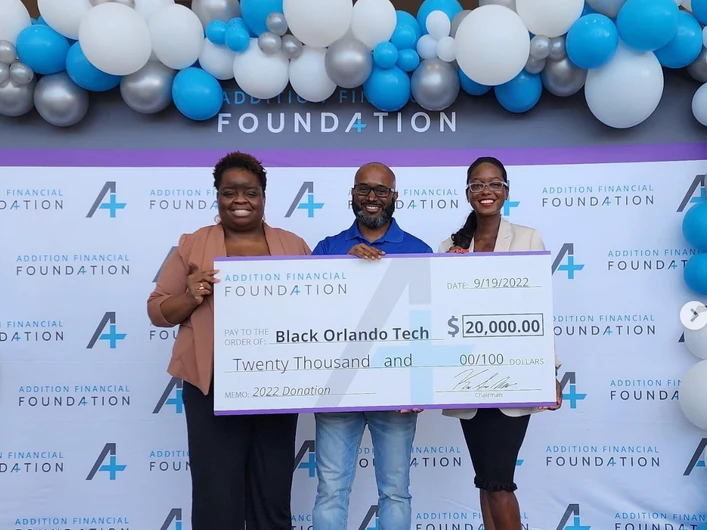 Three people holding an oversized Addition Financial Foundation cheque for $20,000, in front of a balloon arch and a branded backdrop