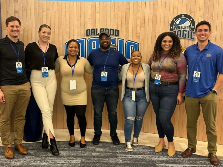 Seven participants wearing lanyards, posed beneath an Orlando Magic sign at the team&rsquo;s innovation challenge