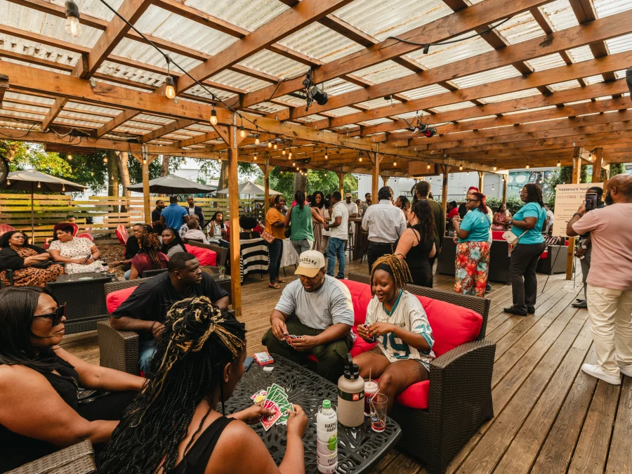 Guests under a string-lit pergola at the Sunset Soir&eacute;e, some playing a card game at a low table while others mingle beside a Sunset Soir&eacute;e banner