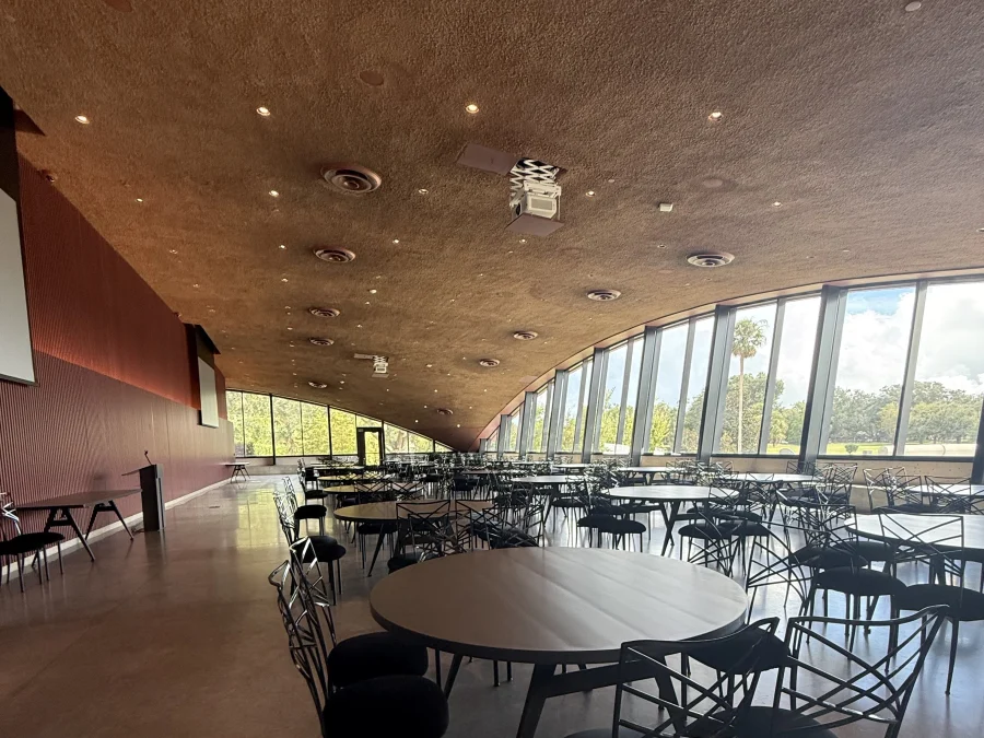 The main hall set with round tables and chairs beneath a curved textured ceiling, with floor-to-ceiling windows looking out over lawns and palms